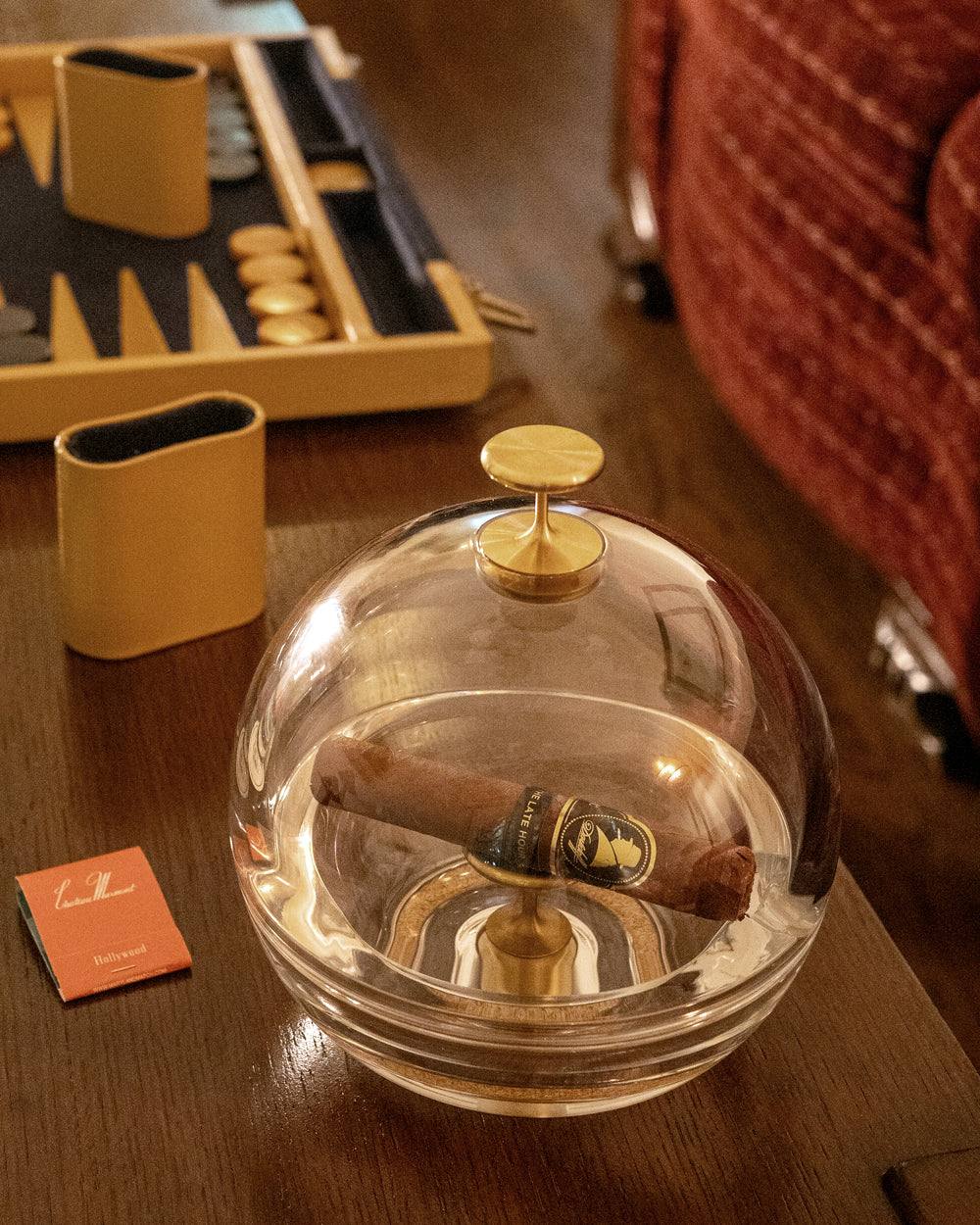 The Murmmr crystal dome closed over a resting cigar on a wooden table, with a backgammon board behind it