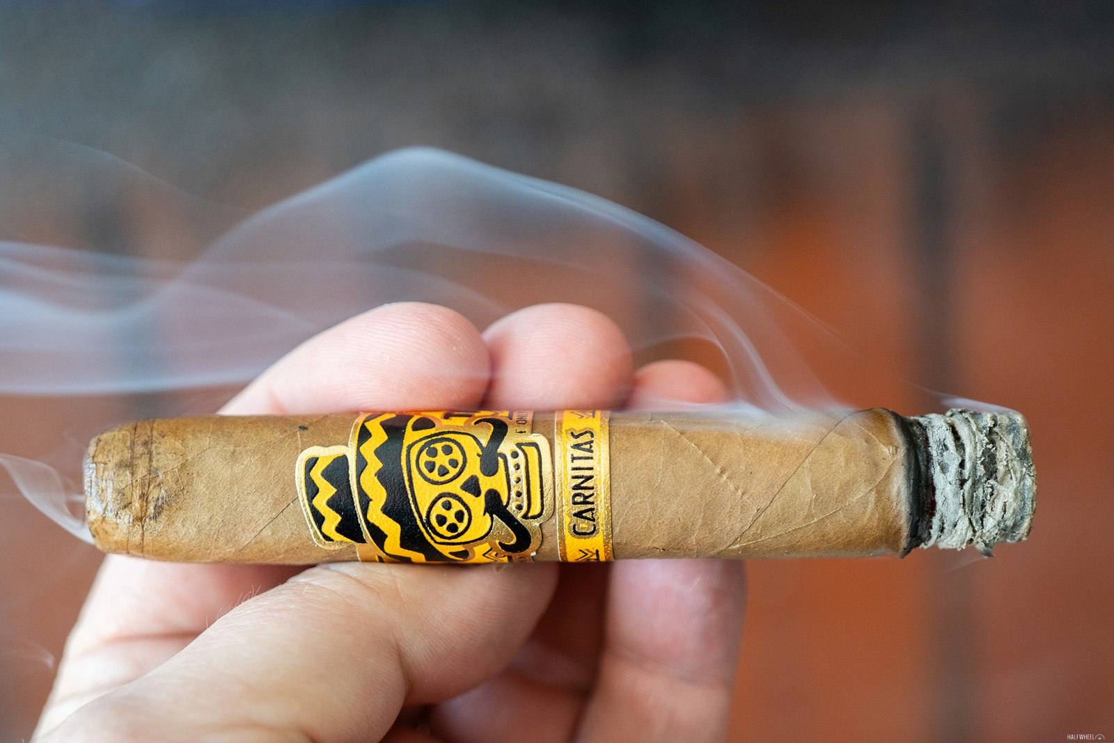 A lit Rojas Street Tacos Carnitas Robusto held in a hand, showing its yellow skull band, photographed by halfwheel